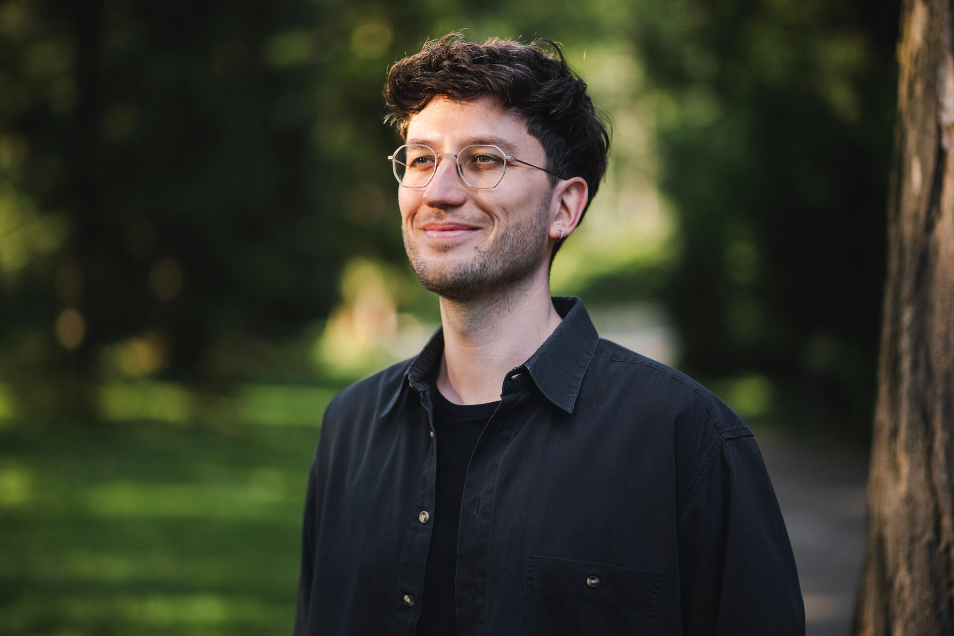 Jakob Bouchal Jakob Bouchal, der ein schwarzes Hemd und eine Brille trägt, steht vor einem Hintergrund satter, grüner Blätter und Pflanzen, und lächelt.