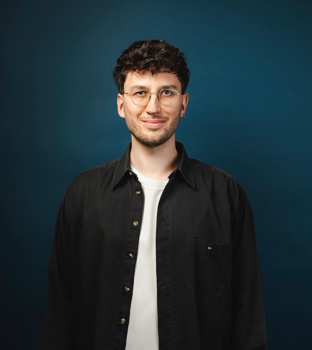 Jakob Bouchal Jakob Bouchal, der ein schwarzes Hemd und ein weißes T-Shirt trägt, steht vor einem dunkleblauen Hintergrund und lächelt.