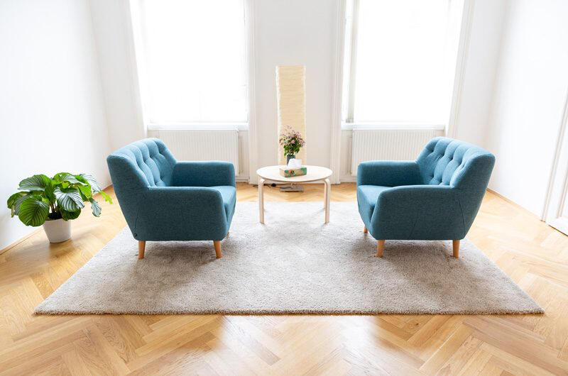 Praxisraum Alserstraße Zwei blaue Polstersessel stehen links und rechts von einem weißen runden Tisch auf einem hellen Teppich. Auf der linken Seite steht eine Pflanze, und in der Mitte des hellen Raums stehen eine hohe Lampe und eine Blumenvase vor großen Fenstern.